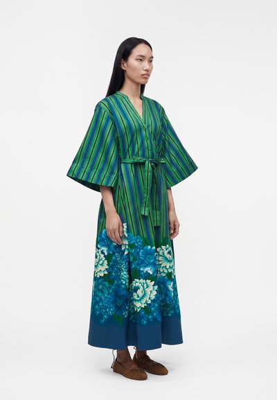 Γυναίκα που φοράει ένα πράσινο και μπλε ριγέ φόρεμα κιμονό με φαρδιά μανίκια, φλοράλ σχέδιο στο τελείωμα και καφέ παπούτσια με κορδόνια.