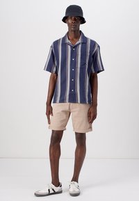 Chemise à rayures bleu marine à manches courtes avec une poche, associée à un short beige. Portée avec un chapeau seau noir et des baskets grises et blanches.