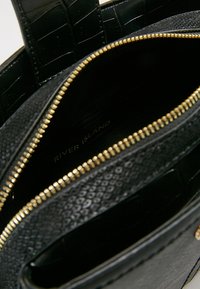 Svart texturerad handväska inuti med en guldfärgad dragkedja, en slät läderbotten och ett präglat mönster på ena sidan. Nitat varumärkesmärke synligt.