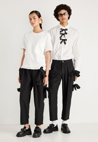 T-shirt branca de algodão com barra انجابا, calças pretas com laços laterais e sapatos pretos com tachas. Estilizado para a moda contemporânea.