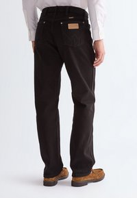 Man wearing black straight-leg jeans with visible Wrangler logo on back pocket, paired with brown suede loafers and white shirt.