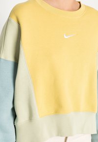 Persoon draagt een gekleurde sweatshirt met gele, lichtgroene en blauwe secties en een klein wit Nike swoosh-logo op de borst.