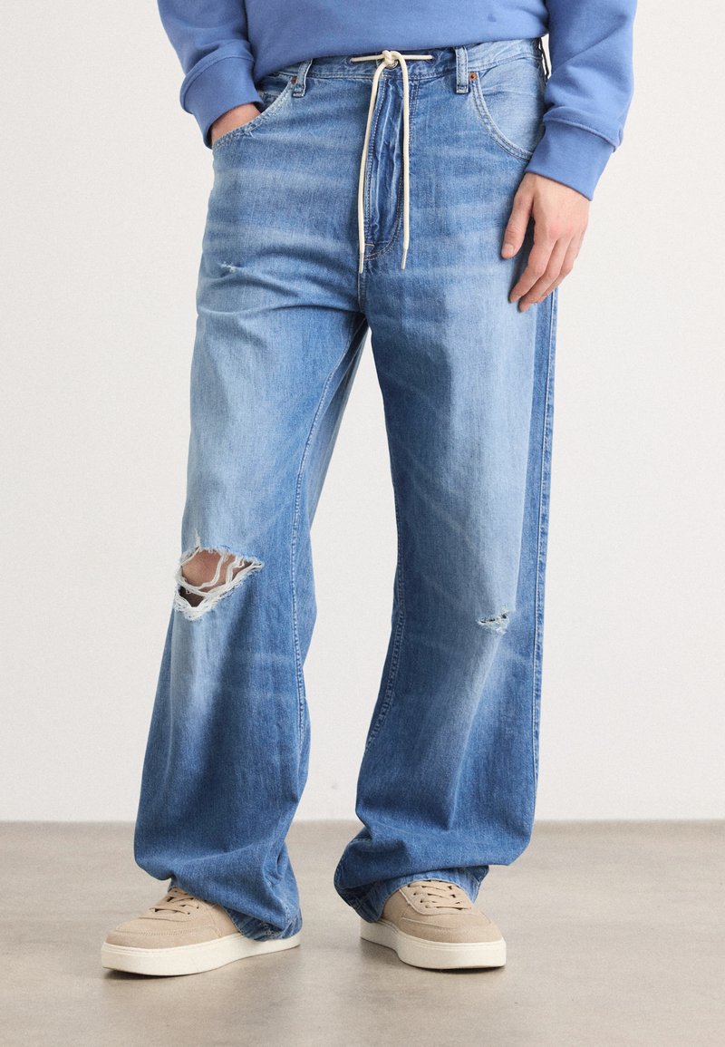 Jeans de ganga de corte largo em azul claro, com detalhes rasgados, featuring uma cintura com cordão e grandes bolsos frontais. Com leve desbotamento.
