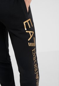 Svarta sweatpants av mjukt tyg med en framträdande guldfärgad tryck med "EA7 Emporio Armani" längs sidan och en sidoficka.