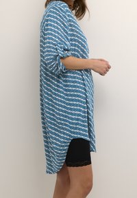 Femme portant une chemise à manches longues bleue à motifs avec des manches retroussées et un short noir avec une bordure en dentelle, se tenant contre un mur blanc.