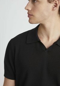 Polo noir à manches courtes en tissu texturé, avec un col classique et un design subtil en V. Esthétique simple et moderne.