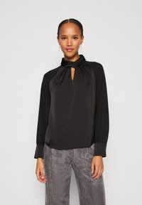 Schwarze Bluse mit einem geknoteten Kragen, verfügt über lange Ärmel und einen glatten, satinähnlichen Stoff. Kombiniert mit grauen strukturierten Hosen.