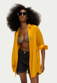 Camicia gialla a bottoni con maniche arrotolate, sopra un top bikini a stampa leopardata. Pantaloncini di denim neri con orlo risvoltato. Occhiali da sole neri a forma quadrata.