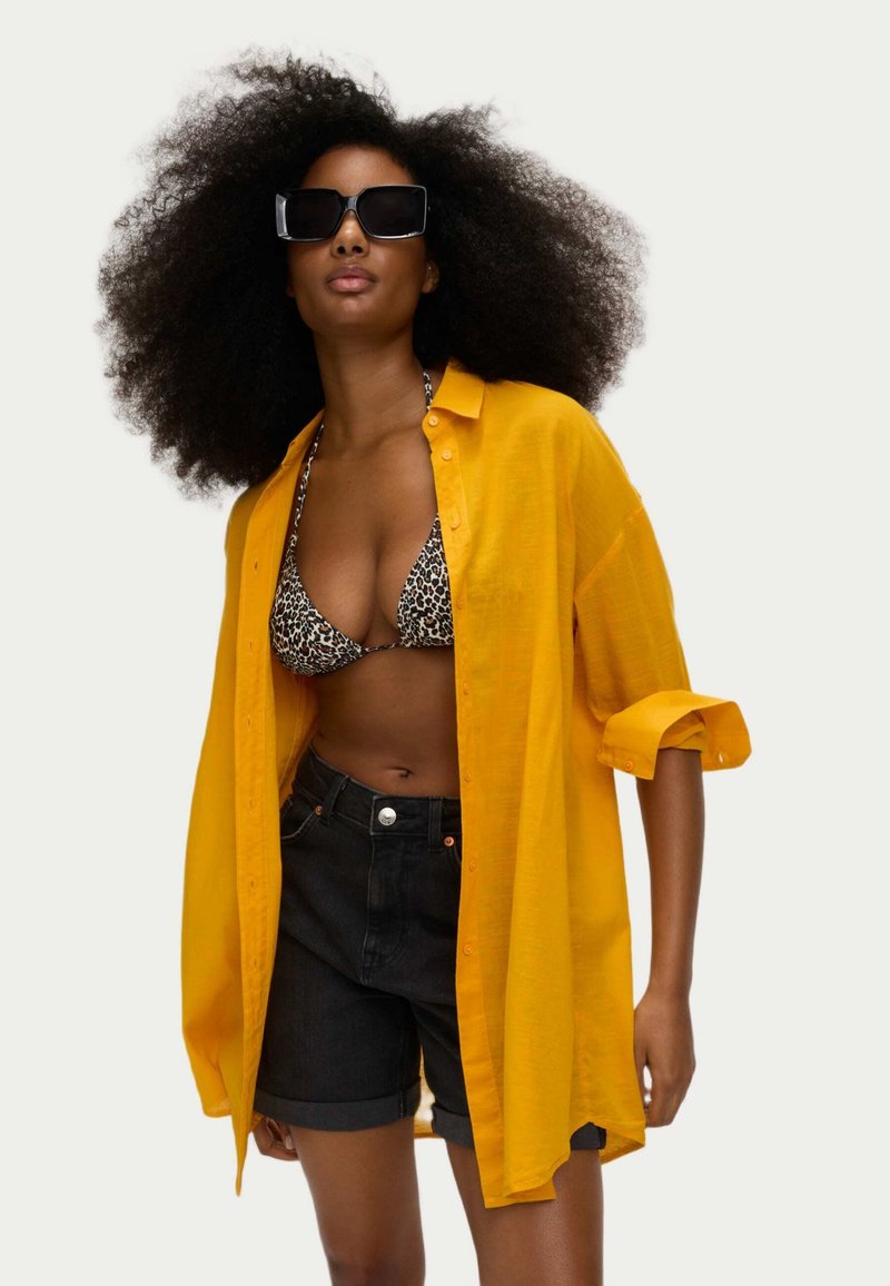 Camicia gialla a bottoni con maniche arrotolate, sopra un top bikini a stampa leopardata. Pantaloncini di denim neri con orlo risvoltato. Occhiali da sole neri a forma quadrata.