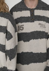 Deux personnes portant des pulls en maille rayés gris et blanc, l'un avec des boutons et le texte imprimé "15" et "MULTI APPAR."