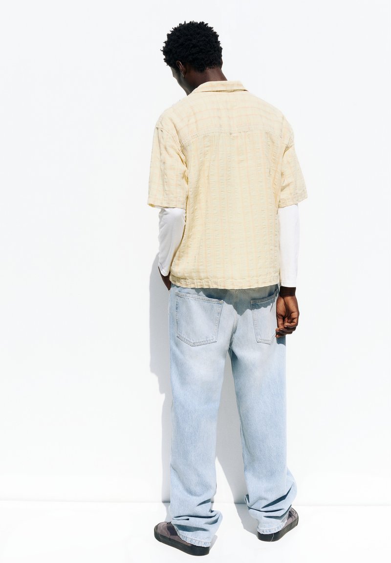 Homme debout face à un mur blanc, portant une chemise jaune clair à manches courtes par-dessus une chemise blanche à manches longues et un jean bleu clair.