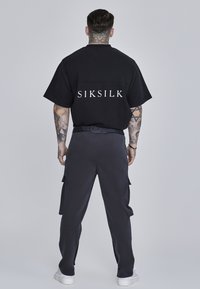 Musta lyhythihainen paita, jossa on "SIKSILK"-logo selässä, yhdessä tummien cargo-housujen ja valkoisten tennareiden kanssa. Sujuva, rento asukokonaisuus.