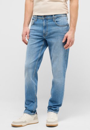 Mann trägt hellblaue Jeans mit geradem Schnitt und weiße Turnschuhe, steht vor einem einfarbig grauen Hintergrund.