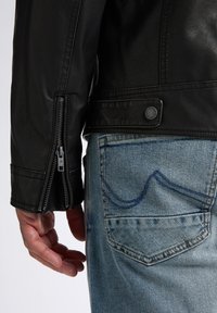 Schwarze Lederjacke mit einem Reißverschluss an den Ärmelbündchen, kombiniert mit hellblauen Jeans, die ein markantes Design der Gesäßtasche aufweisen.