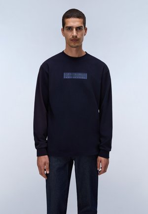 Uomo che indossa una felpa blu navy con un piccolo logo frontale e jeans scuri, in piedi davanti a uno sfondo chiaro e semplice.