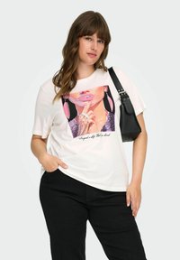 Valkoinen t-paita, jossa on värikäs graafinen printti naisen kasvoista vaaleanpunaisilla asusteilla, yhdistettynä mustiin housuihin ja pieneen mustaan laukkuun.