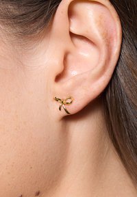 Złote kolczyki w kształcie kokardy, z gładkimi powierzchniami i błyszczącym wykończeniem, z małym centralnym akcentem.