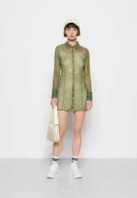 Vestido camiseiro transparente verde com padrão, mangas longas e colarinho, combinado com um boné amarelo claro, mala creme e ténis brancos.