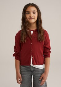 Nuori tyttö, jolla on pitkät ruskeat hiukset, pukeutunut punaisiin nappikauluksellisiin neuletakkeihin valkoisen paidan ja harmaiden farkkujen kanssa neutraalia taustaa vasten.