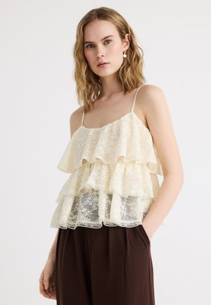 Mujer joven que lleva una blusa sin mangas de encaje en crema con volantes y pantalones marrón oscuro, de pie con una mano en el bolsillo contra un fondo blanco.
