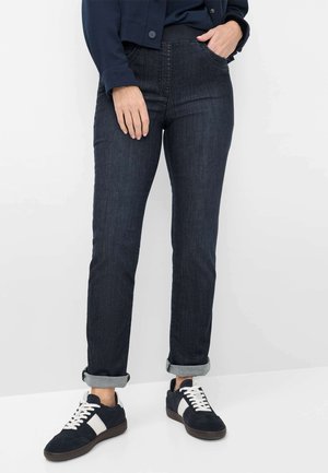 Person trägt dunkel hochgekrempelte Jeans, marineblaue Jacke und schwarz-weiße Sneaker, steht vor einem hellgrauen Hintergrund.