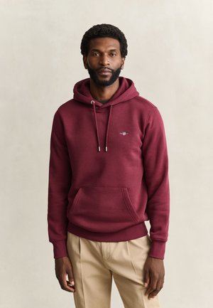 Mann mit kurzen lockigen Haaren und Bart, der einen bordeauxfarbenen Hoodie und beige Hosen trägt und vor einem einfarbigen hellen Hintergrund steht.