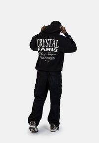 Felpa nera con testo bianco c "CRYSTAL PARIS Jewelry & Timepieces" e "A RICH PARENTS" sul retro. Abbinata a pantaloni cargo neri e sneakers.