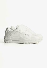 White sneakers made of smooth material, featuring a low profile, padded collar, flat laces, and textured outsole. Simple design with minimal accents.
