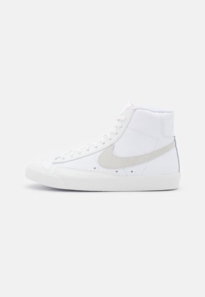 Baskets montantes blanches avec logo Nike Swoosh gris clair, fermeture à lacets et semelle en caoutchouc, présentées en profil latéral sur fond uni.