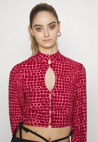 Blusa com padrão vermelho, detalhe em recorte em forma de fechadura, mangas longas e gola alta. Apresenta fechos de botão e um design cropped. Textura suave.