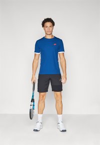 T-shirt de sport bleu avec des accents blancs, associé à un short noir. Le modèle tient une raquette de tennis, portant des chaussettes et des chaussures de sport blanches.