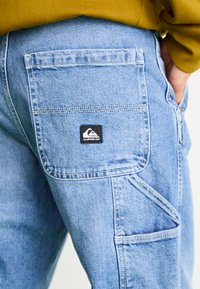 Light blue denim cargo pants featuring a visible black Quiksilver logo patch on the back pocket, with a sturdy, textured fabric and multiple pockets.
