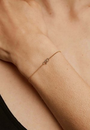 Pulsera de cadena de oro con un charm minimalista en forma de clip de papel, textura suave y eslabones finos, llevada en una muñeca sobre un fondo neutro.