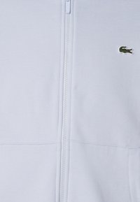 Vêtement blanc à fermeture éclair centrale avec petit logo crocodile vert en haut à droite.