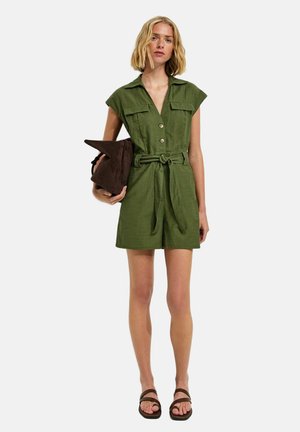 Femme portant une combinaison short à manches courtes olive avec ceinture, tenant une pochette marron foncé, portant des sandales marron, debout devant un fond blanc.