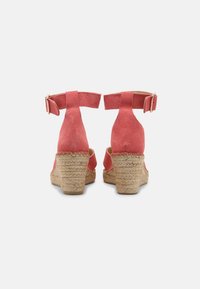 Un paio di sandali con zeppa in suede corallo, dotati di cinturini alla caviglia e tacco in juta intrecciata, visti da dietro su uno sfondo bianco.