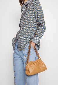 Borsa in pelle marrone chiaro con parte superiore arricciata e manico in catena color marrone chiaro, accompagnata da una camicia a fantasia blu e marrone e pantaloni buttoned blu chiaro.