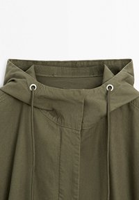 Olivgrüne Kapuzenjacke mit Kordelzügen, die durch Metallösen geführt sind, und sichtbaren Steppnähten am Kragen und vorne.