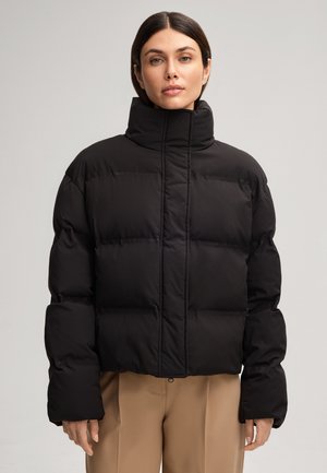 Veste rembourrée noire avec un col montant, fermeture éclair et grands motifs en boudin. Texture lisse et mate ; matériau léger.