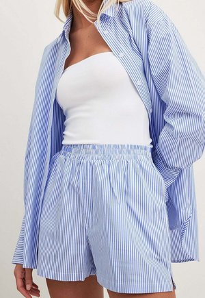 Vrouw met een blouse met verticale blauw-witte strepen en bijpassende short, een witte tube top, hand in zak, tegen een effen achtergrond.