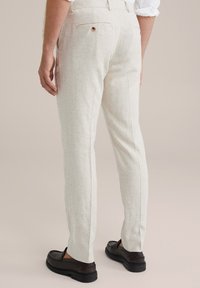 Világos bézs vászon nadrág, testhezálló fazonnal, hátsó zsebekkel és egy gombos részlettel. Fekete lakkcipővel viselve.