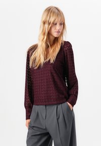 Blonde vrouw in een gestructureerde bordeauxrode blouse met lange mouwen en losse grijze broek, poseert met één hand in haar zak tegen een witte achtergrond.