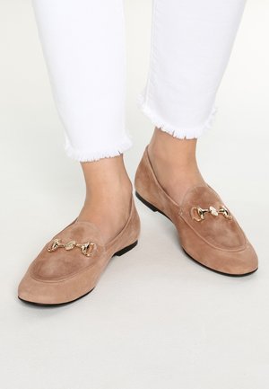 Lys beige suede loafers med rund tå, guld kæde accent på toppen, og flad sort sål, parret med hvide jeans med flosset kant.