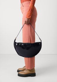 Black, crescent-shaped handbag with a glossy finish and zipper closure, held by a person wearing light pink pants and brown lace-up boots.