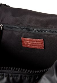 Černá látková taška s hladkým interiérem; obsahuje zipovou kapsu a hnědou koženou nálepku s vyraženým nápisem "United Colors of Benetton."