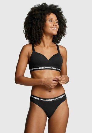 Frau mit lockigem Haar, die einen schwarzen BH und Unterwäsche der Marke Puma trägt, lächelt und schaut seitlich vor einem schlichten Hintergrund.