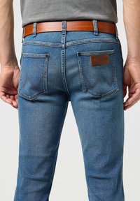 Jeans en denim bleu avec une coupe slim, présentant deux poches arrière avec des coutures signature et une étiquette en cuir marron. Une ceinture marron améliore le look.