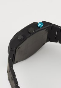 Montre noire avec un cadran carré, bracelet en métal, boutons ronds et une touche de bleu. Fini mat avec des détails en quincaillerie métallique.