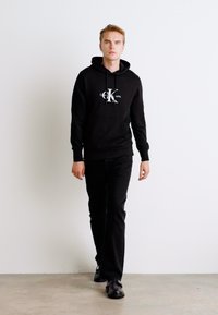 Felpa nera con logo frontale, abbinata a pantaloni neri e scarpe. Il modello cammina su un pavimento in cemento chiaro contro uno sfondo neutro.