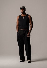 Chemise sans manches noire, pantalon noir, ceinture brune texturée et chaussures noires avec des accents crème. Les accessoires comprennent des colliers superposés et une casquette.